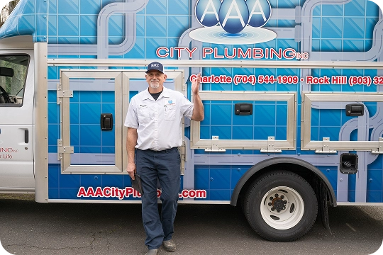 A man in a AAA City Plumbing uniform stands next to a company service van, smiling and holding the door handle.